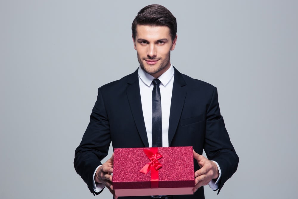 Handsome businessman holding gift box over gray background and looking at camera
